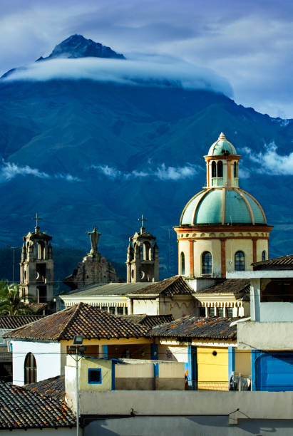 A cloud surrounds the peak of the Cotacachi Volcano, a dormant volcano in the northern Ecuadorian Andes. The mountain provides a dramatic backdrop for the Spanish colonial dome of the Iglesia El Jordan in the market town of Otavalo.